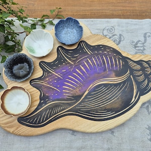 Könnte beinhalten: Ein helles Holz-Servierbrett in Form einer Muschel mit einer großen, dunkel umrandeten violett-schwarzen Muschelgrafik. Vier kleine, unterschiedlich gefärbte Keramikschalen sind auf der Oberfläche des Brettes angeordnet.