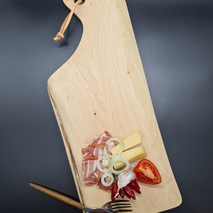 Könnte beinhalten: Ein Holzbrett mit Griff, auf dem eine Auswahl an getrocknetem Fleisch, Käse, Zwiebelringen, Knoblauch und roten Paprikaschoten liegt. Zwei silberne Besteckteile liegen auf dem Brett.