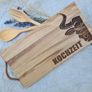 May include: A wooden cutting board with a bull's head design and the word "KOCHZEIT" engraved on it. The board has a handle and is made of light brown wood.