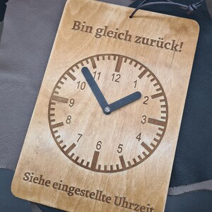 Op de afbeelding: Een houten bord met de tekst "Bin gleich zurück!" en een klok met zwarte wijzers. De tekst "Siehe eingestellte Uhrzeit" staat onderaan het bord.