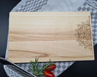 May include: A rectangular wooden cutting board with a mandala design etched into the corner. The board is light brown with visible wood grain. Rosemary sprigs and cherry tomatoes are placed near a knife on a checkered cloth.