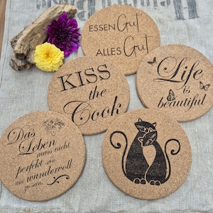 May include: A set of six round cork coasters. Each coaster has a unique design, including phrases like "Kiss the Cook" and "Life is Beautiful," and a black cat silhouette. The coasters are a natural cork color.
