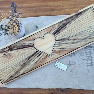 Könnte beinhalten: Ein Holzbrett mit einer Herzform, die mit String Art erstellt wurde. Das Herz ist aus schwarzem Garn gefertigt und der Hintergrund ist aus hellem braunem Holz.