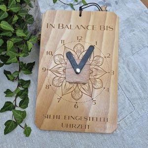May include: A rectangular wooden wall clock featuring a laser-engraved mandala design and the text IN BALANCE BIS SIEHE EINGESTELLTE UHRZEIT. The clock has two dark, thick hands attached to a central square wooden piece on a light wood grain board.