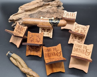 May include: Wooden cigar rests with various engraved sayings, including "Cigars Are Always" and "Time To Drink &". The rests are made of light and dark wood and have intricate designs.
