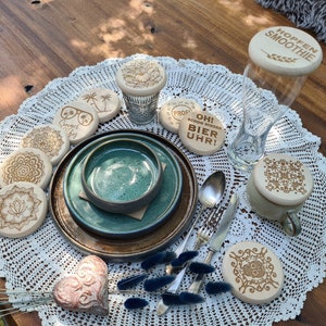 Op de afbeelding: Een set houten onderzetters met verschillende ontwerpen en spreuken, waaronder "Summer Vibes", "Hopfen Smoothie", "Oh! Schon Wieder Bier Uhr!" en "Prost!" De onderzetters zijn gerangschikt op een wit kantkleed met een teal keramische kom en een bestekset.