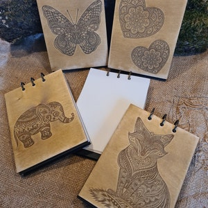 Può includere: Quaderni in legno con disegni incisi al laser di una farfalla, cuori, un elefante e una volpe. Ogni quaderno ha una rilegatura a spirale nera e pagine bianche vuote. Il legno è di colore chiaro.