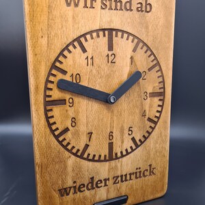 Op de afbeelding: Een houten klok met een zwart wijzerplaat en wijzers. De wijzerplaat heeft de cijfers 1 tot en met 12 en de tekst "Wir sind ab wieder zurück".