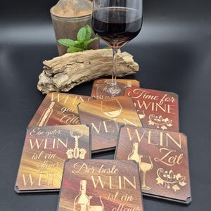 Può includere: Una collezione di sottobicchieri in legno con disegni a tema vino e testo in tedesco. I sottobicchieri presentano immagini di bottiglie di vino, bicchieri e cavatappi. Un bicchiere di vino rosso è in primo piano, con un contenitore di metallo rustico e legno sullo sfondo.