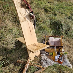Può includere: Una sedia di legno con un design di cervo intagliato e una cinghia di cuoio. La sedia è seduta su una superficie erbosa con una cassa di legno e bottiglie di liquido.