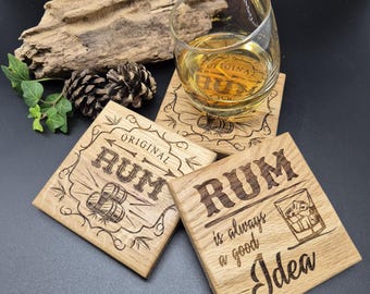 May include: Four wooden coasters with a rum theme. The coasters are square and have a light brown wood finish. The top of each coaster has a different design, including the words "Original Rum", "Rum is always a good idea", and a glass of rum with ice.