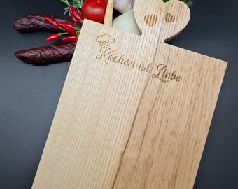 May include: A wooden cutting board with a heart-shaped handle and the text "Kochen ist Liebe" (Cooking is Love) engraved on the surface. The board is surrounded by vegetables and herbs, including a red tomato, green onions, garlic, and red chili peppers.
