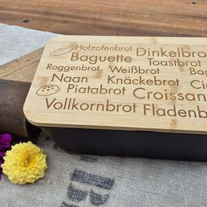 Op de afbeelding: Een zwarte brooddoos met een bamboe deksel. Het deksel is gegraveerd met verschillende broodnamen en illustraties. Paarse en gele bloemen en een metalen voorwerp liggen naast de brooddoos.
