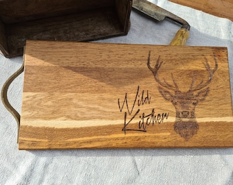 May include: A wooden cutting board with a leather handle. The board is engraved with a stag and the words "Wild Kitchen".