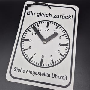 Op de afbeelding: Wit bord met zwarte tekst en een klok. Het bord staat "Bin gleich zurück!" en "Siehe eingestellte Uhrzeit".