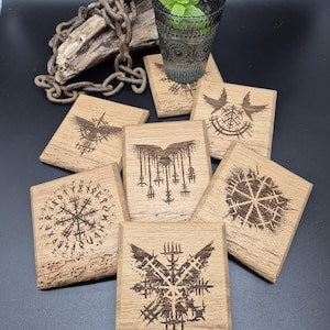 Puede incluir: Conjunto de seis posavasos de madera con varios símbolos y diseños nórdicos, incluyendo un cuervo, un copo de nieve y un círculo rúnico. Los posavasos están hechos de madera marrón claro y tienen un aspecto rústico y artesanal.
