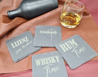 May include: A collection of slate coasters with white lettering, including "Whisky Time" and "Rum Time." A black bottle and a glass of amber liquid are also present. The coasters are square and rest on a brown leather surface.