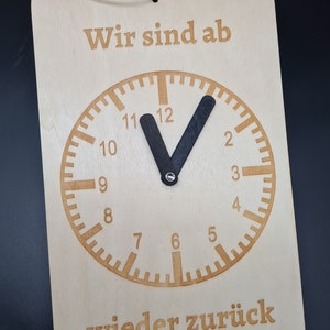 Op de afbeelding: Een houten bord met de tekst "Wir sind ab wieder zurück" en een klok met zwarte wijzers die op 12:00 uur wijzen.