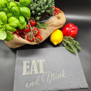 May include: A slate serving board with the text "EAT and Drink" in white lettering. The board is surrounded by fresh herbs, tomatoes, peppers, and a lemon.