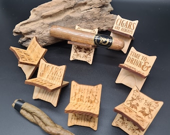 May include: Six wooden cigar rests with various engraved sayings, including "Cigars are always in style" and "Time to drink". One rest has a cigar resting on it. A brown cigar is lying on a dark surface.