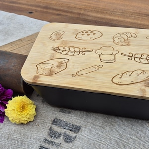 May include: A black rectangular bread pan with a bamboo lid. The lid is engraved with various bread-related illustrations. A rusty metal object, wooden plank, and purple and yellow flowers are next to the pan.