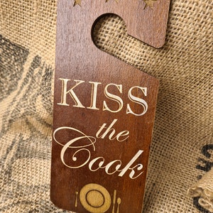 Op de afbeelding: Een houten deurhanger met een bruine afwerking en een witte inscriptie die "KISS the Cook" luidt. De hanger heeft een haak aan de bovenkant en een ontwerp van een bord en bestek aan de onderkant.