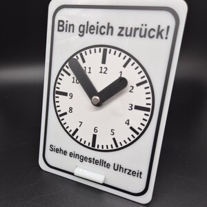 Op de afbeelding: Een wit bord met zwarte tekst en een klok. De tekst luidt "Bin gleich zurück!" wat "Ik ben zo terug!" betekent en "Siehe eingestellte Uhrzeit" wat "Zie de ingestelde tijd" betekent.