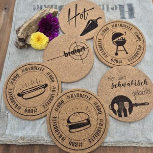 Op de afbeelding: Een verzameling ronde kurken onderzetters met zwarte opdrukken en tekst. Ontwerpen omvatten een chilipeper, een grill, een hotdog, een hamburger en het woord "Hot". De onderzetters zijn voor BBQ en grillen.