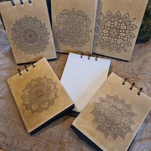 Pode incluir: Cinco pequenos cadernos marrons com espiral e capas pretas. Cada caderno apresenta um design de mandala intrincado diferente em preto.