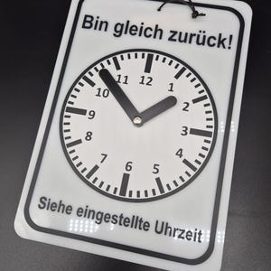 Op de afbeelding: Een wit bord met zwarte tekst en een klok. Het bord staat "Bin gleich zurück!" wat "Ik ben zo terug!" betekent en "Siehe eingestellte Uhrzeit" wat "Zie de ingestelde tijd" betekent.