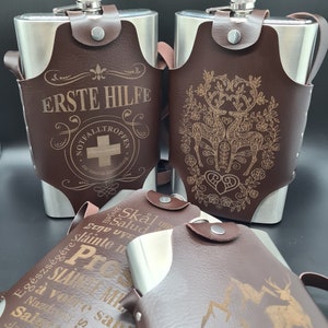 May include: Three brown leather-wrapped stainless steel hip flasks with different engraved designs. The first flask has the text "Erste Hilfe" and a red cross. The second flask has a design of two deer with a heart. The third flask has a mountain scene with a deer.