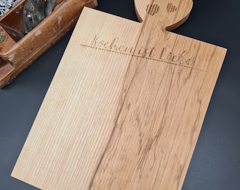 May include: A wooden cutting board with a heart-shaped handle. The board has a light brown color and a natural wood grain. The text "Kochen ist Liebe" is engraved on the board.