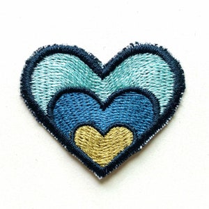Puede incluir: Un parche bordado en forma de corazón azul y dorado con tres capas de corazones. La capa exterior es azul claro, la capa intermedia es azul oscuro y la capa interior es dorada.
