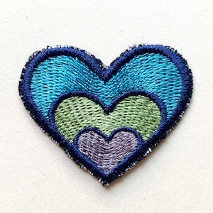 Könnte beinhalten: Ein gestickter Herz-Patch in Blau, Grün und Lila. Das Herz besteht aus drei konzentrischen Herzen, wobei das kleinste Herz lila, das mittlere Herz grün und das größte Herz blau ist.