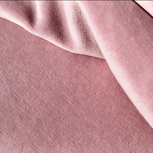 May include: Close-up of a soft, pink velvet fabric with a subtle texture. The fabric is folded over itself, creating a smooth, flowing drape.