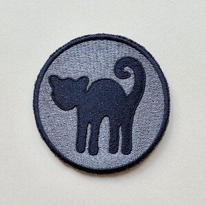 Op de afbeelding: Een grijze ronde patch met een geborduurd silhouet van een zwarte kat.