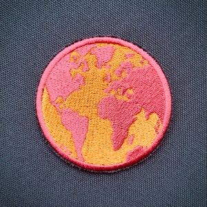 Może przedstawiać: Naszywka z haftowanym obrazem mapy świata w kolorze różowym i pomarańczowym. Naszywka jest okrągła i ma różową obwódkę.