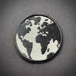 Könnte beinhalten: Ein gestickter schwarzer und weißer Aufnäher der Erde mit einem weißen Umriss. Der Aufnäher ist rund und hat einen schwarzen Rand.
