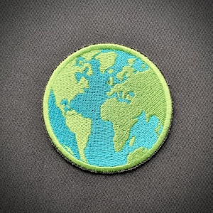 Puede incluir: Un parche bordado de la Tierra en verde y azul, sobre un fondo de tela gris.