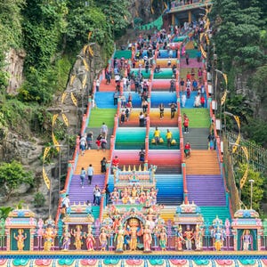 May include: A vibrant, colorful staircase leading up to a temple, with each step painted a different color of the rainbow. People are seen walking up and down the stairs. The temple is surrounded by lush green trees and rock formations.
