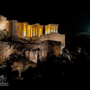 May include: A night view of the Acropolis in Athens, Greece, illuminated by spotlights. The full moon is visible in the sky above the ancient ruins.