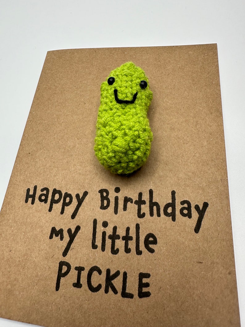 May include: A brown card with the text "Happy Birthday my little PICKLE" written in black marker. A green crocheted pickle with a smiling face is attached to the top of the card.