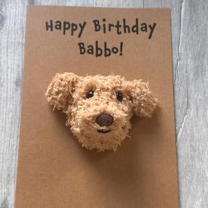 Könnte beinhalten: Ein beiges, flauschiges, dreidimensionales Hunde-Applikations-Gesicht auf einer rechteckigen braunen Karte. Der Text "Happy Birthday Babbo!" ist in schwarzer Tinte über dem Hund gedruckt. Der Hund hat dunkle Augen, eine braune Nase und einen kleinen Mund.
