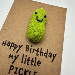 May include: A brown card with the text "Happy Birthday my little PICKLE" written in black marker. A green crocheted pickle with a smiling face is attached to the card.