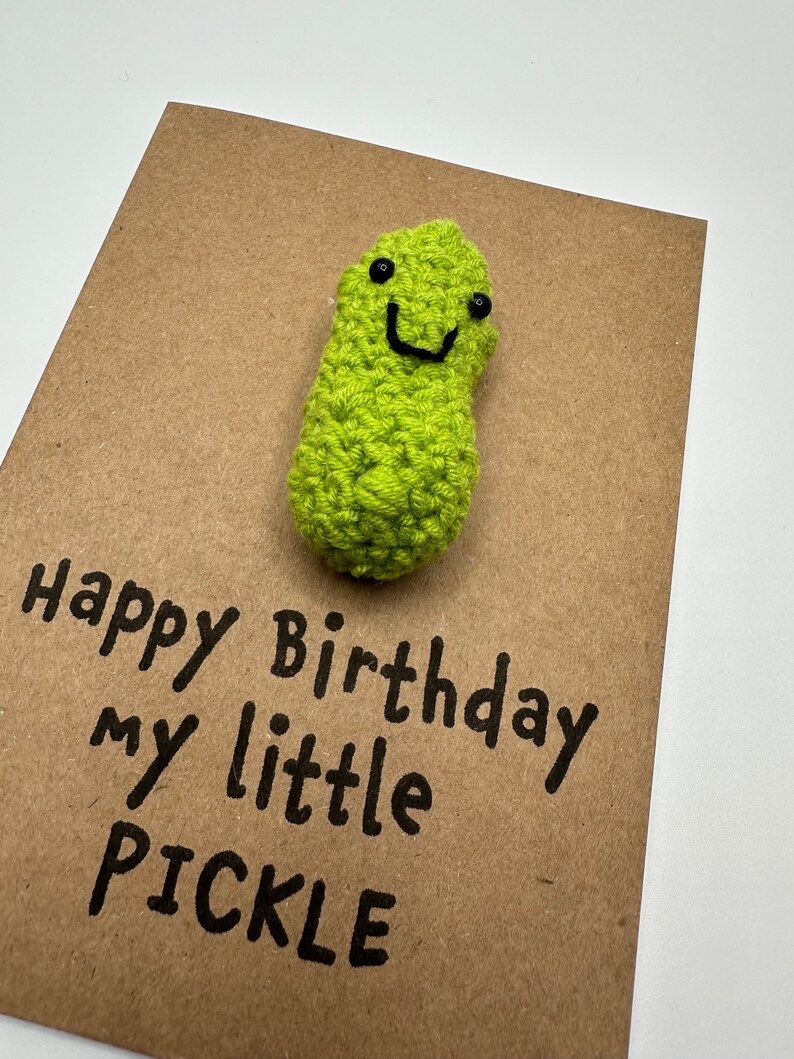 May include: A brown card with the text "Happy Birthday my little PICKLE" written in black marker. A green crocheted pickle with a smiling face is attached to the card.