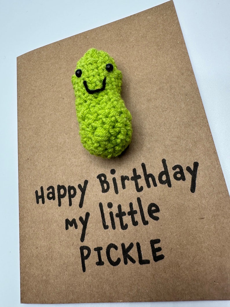 May include: A brown card with the text "Happy birthday my little pickle" written in black marker. A green crocheted pickle with a smiling face is attached to the card.