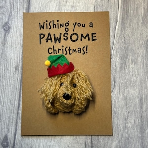 May include: A brown Christmas card featuring a crocheted dog wearing an elf hat. The card reads "Wishing you a PAWSOME Christmas!". The dog is light brown with a black nose and the elf hat is red and green.