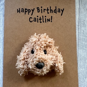 May include: A brown kraft paper greeting card with the black printed text Happy Birthday Caitlin! featuring a three-dimensional, fuzzy, light tan yarn dog head attached to the center with two black eyes and a black nose.
