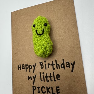 May include: A brown card with the text "Happy Birthday my little PICKLE" written in black marker. A green crocheted pickle with a smiling face is attached to the card.