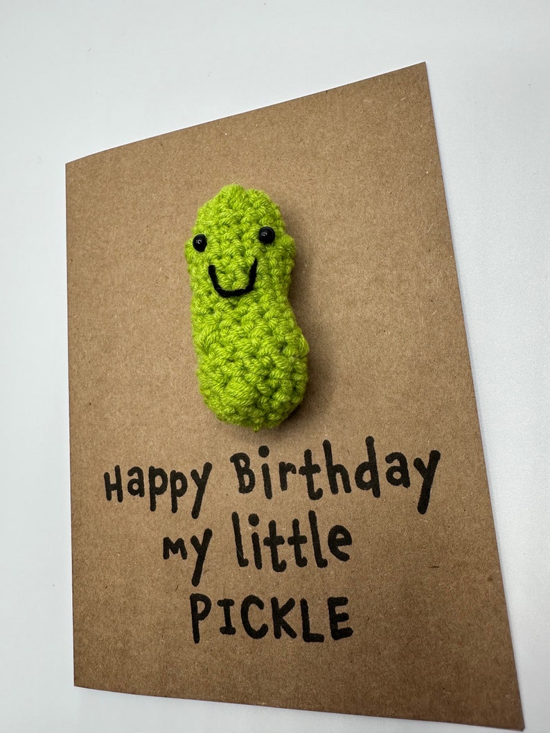 May include: A brown card with the text "Happy Birthday my little PICKLE" written in black marker. A green crocheted pickle with a smiling face is attached to the card.